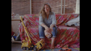 Image of a woman in denim sitting on a couch covered with a red-patterned cover. A small bulldozer lays at her feet.