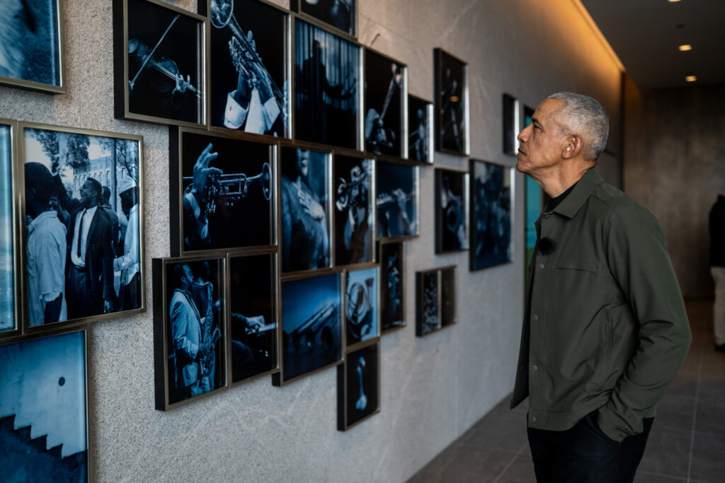 President Barack Obama stands in profile inside the Obama Presidential Center, viewing Carrie Mae Weems’ The Cool Blue Wind. The installation features a grid of blue-toned photographic images mounted along a wall, including scenes of jazz musicians and moments associated with Obama’s 2008 presidential campaign.
