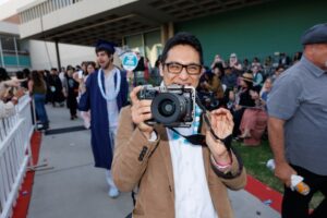 A CalArts student holds a camera in shooting position while smiling during the CalArts graduation march.