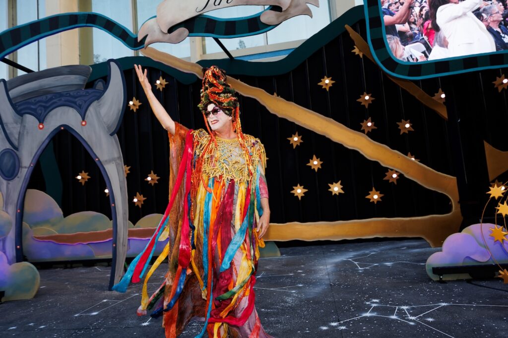 A person in a colorful costume stands on stage at a graduation event with a decorative starry backdrop.