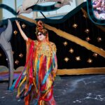 A person in a colorful costume stands on stage at a graduation event with a decorative starry backdrop.