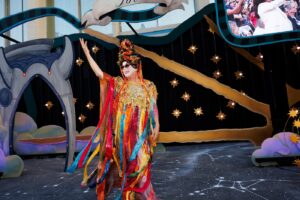 A person in a colorful costume stands on stage at a graduation event with a decorative starry backdrop.
