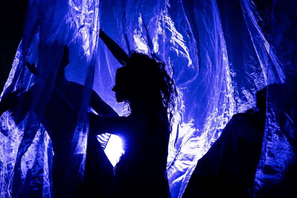 Three silhouetted performers interact with translucent blue fabric, illuminated by soft light, creating an ethereal atmosphere.