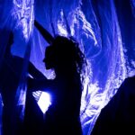 Three silhouetted performers interact with translucent blue fabric, illuminated by soft light, creating an ethereal atmosphere.
