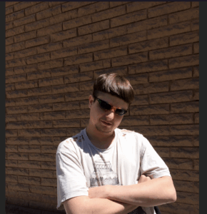 oliver tree in a t-shirt, shades, arms crossed against a brick wall.