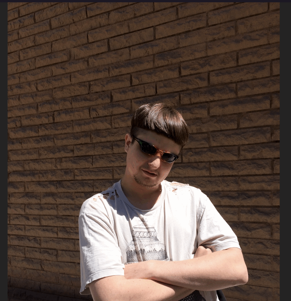oliver tree in a t-shirt, shades, arms crossed against a brick wall.