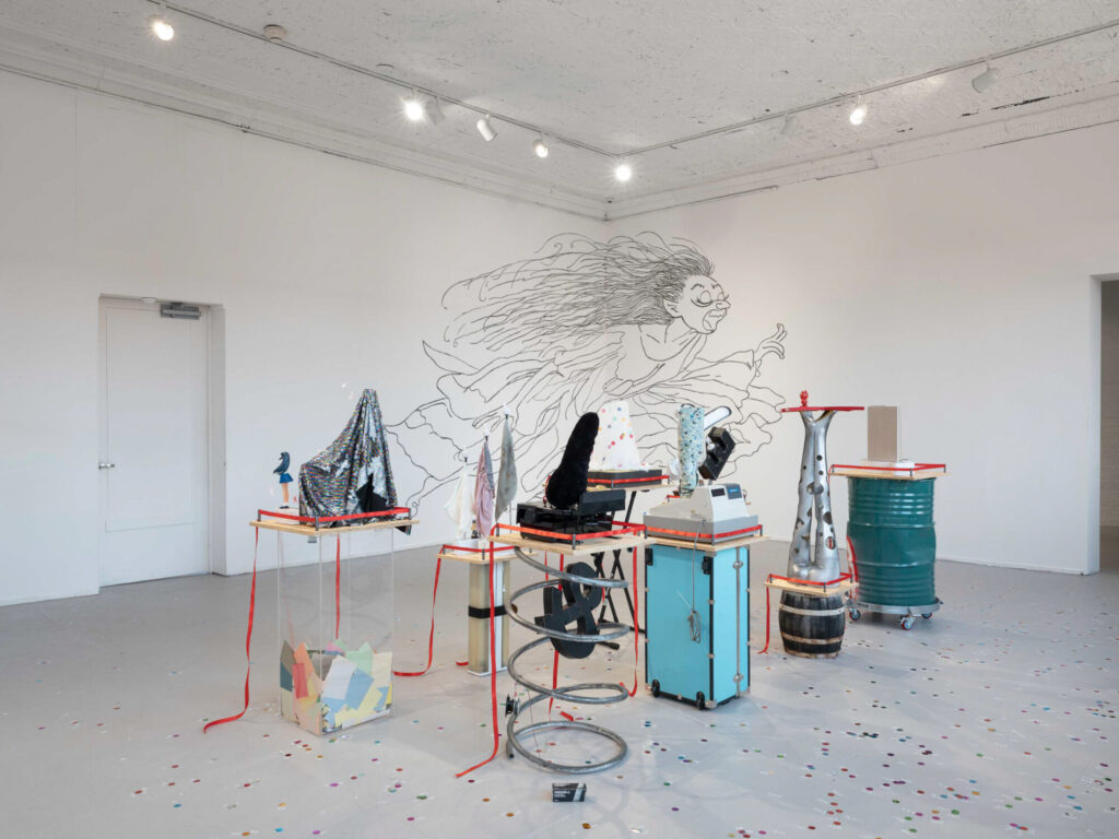 An art exhibition installation featuring various sculptures on pedestals, a whimsical mural on the wall, and colorful confetti on the floor.