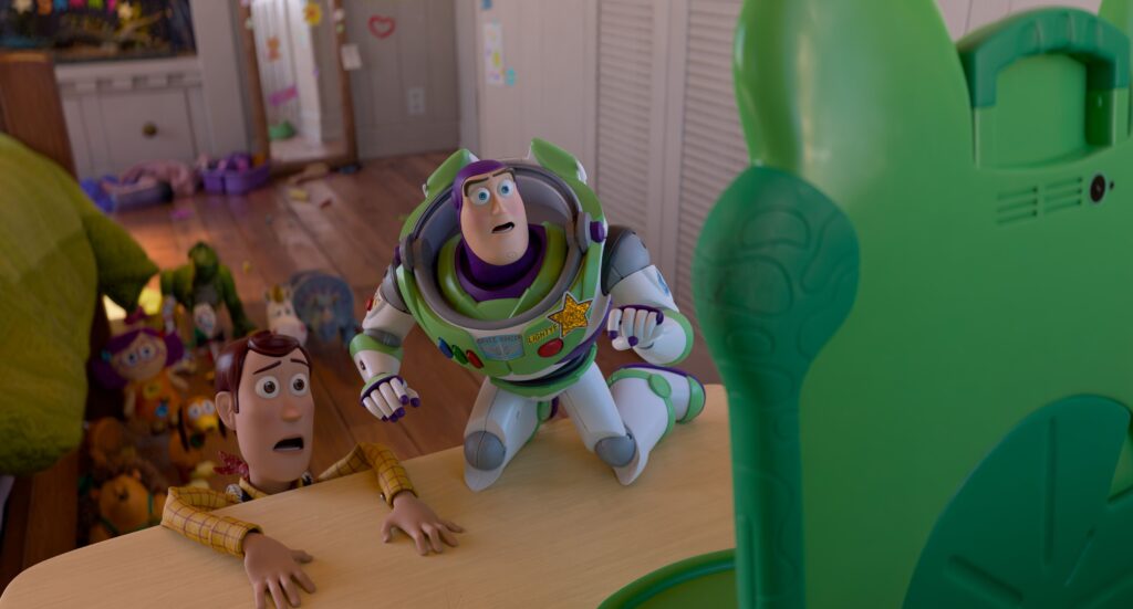 The image from Toy Story 5 shows two animated toy characters on a light wooden table in a child's playroom. The character on the left, Woody, is dressed in a cowboy outfit with a yellow and red theme, appears alarmed as it clings to the table's edge. Buzz Lightyear stands next to Woody.