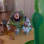 The image from Toy Story 5 shows two animated toy characters on a light wooden table in a child's playroom. The character on the left, Woody, is dressed in a cowboy outfit with a yellow and red theme, appears alarmed as it clings to the table's edge. Buzz Lightyear stands next to Woody.
