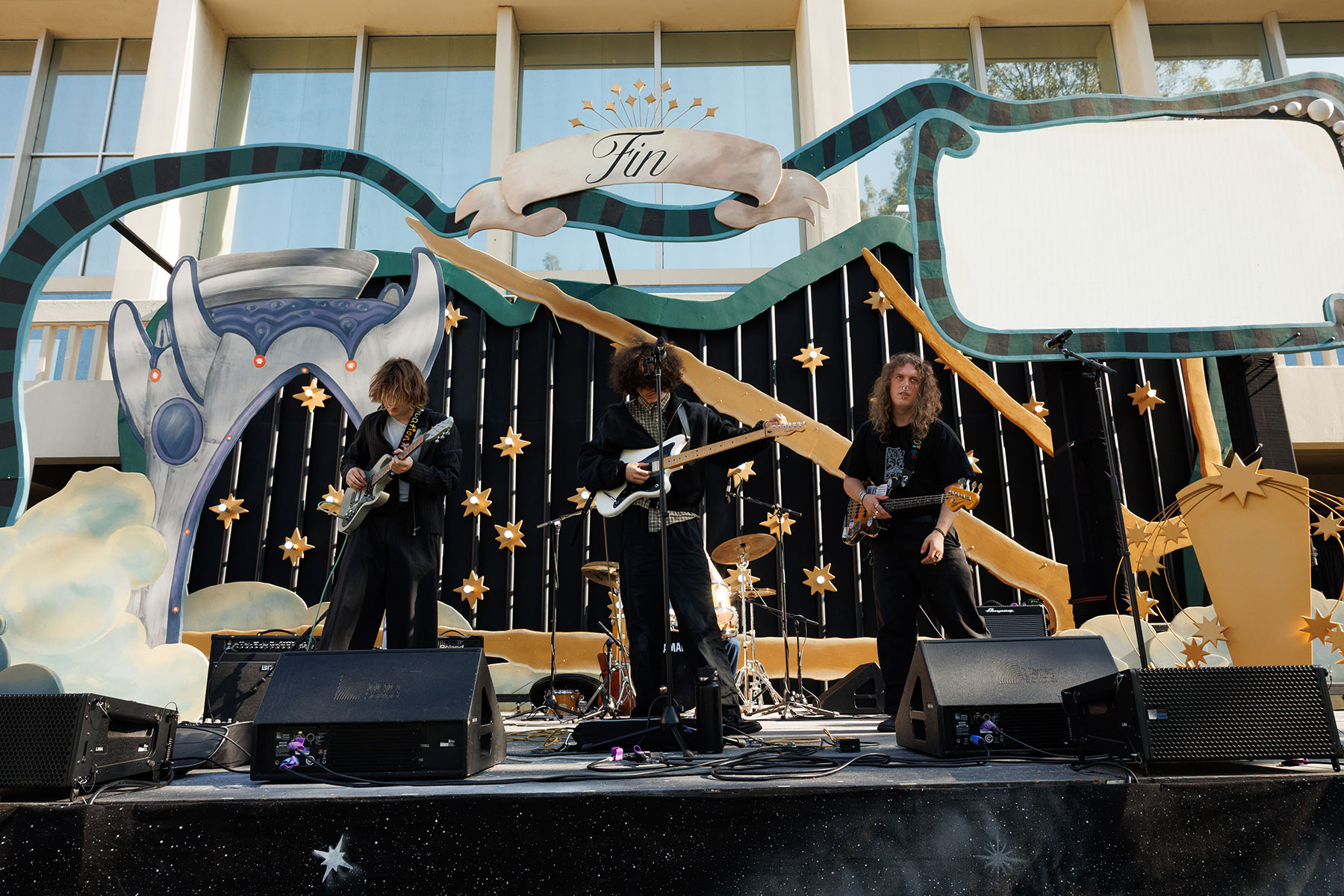Band performing on an outdoor stage with decorative star and cloud-like elements.