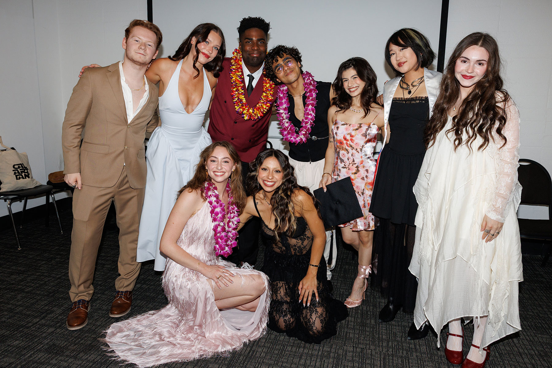 A group of eight people in formal attire, some wearing leis, in a room with plain walls and a carpeted floor.