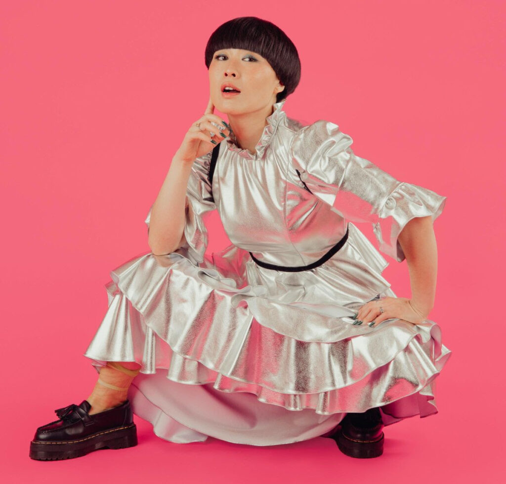 Atsuko Okatsuka crouching in a silver dress with hand on her chin against a pink backdrop.