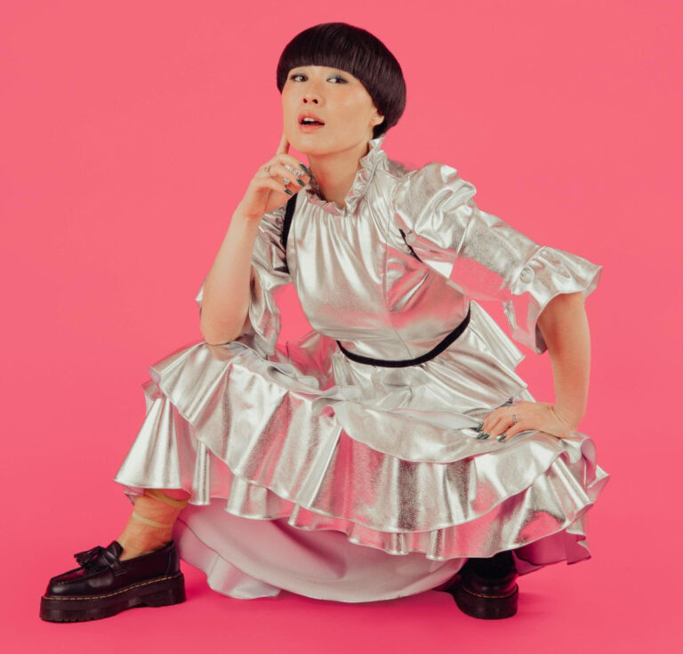 Atsuko Okatsuka crouching in a silver dress with hand on her chin against a pink backdrop.