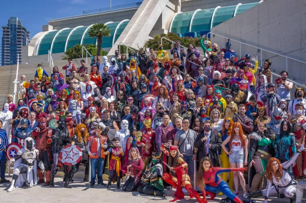 A diverse group of people dressed in various superhero costumes stands on steps outdoors, with modern architecture in the background.