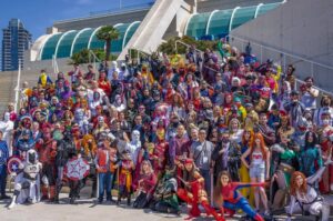 A diverse group of people dressed in various superhero costumes stands on steps outdoors, with modern architecture in the background.
