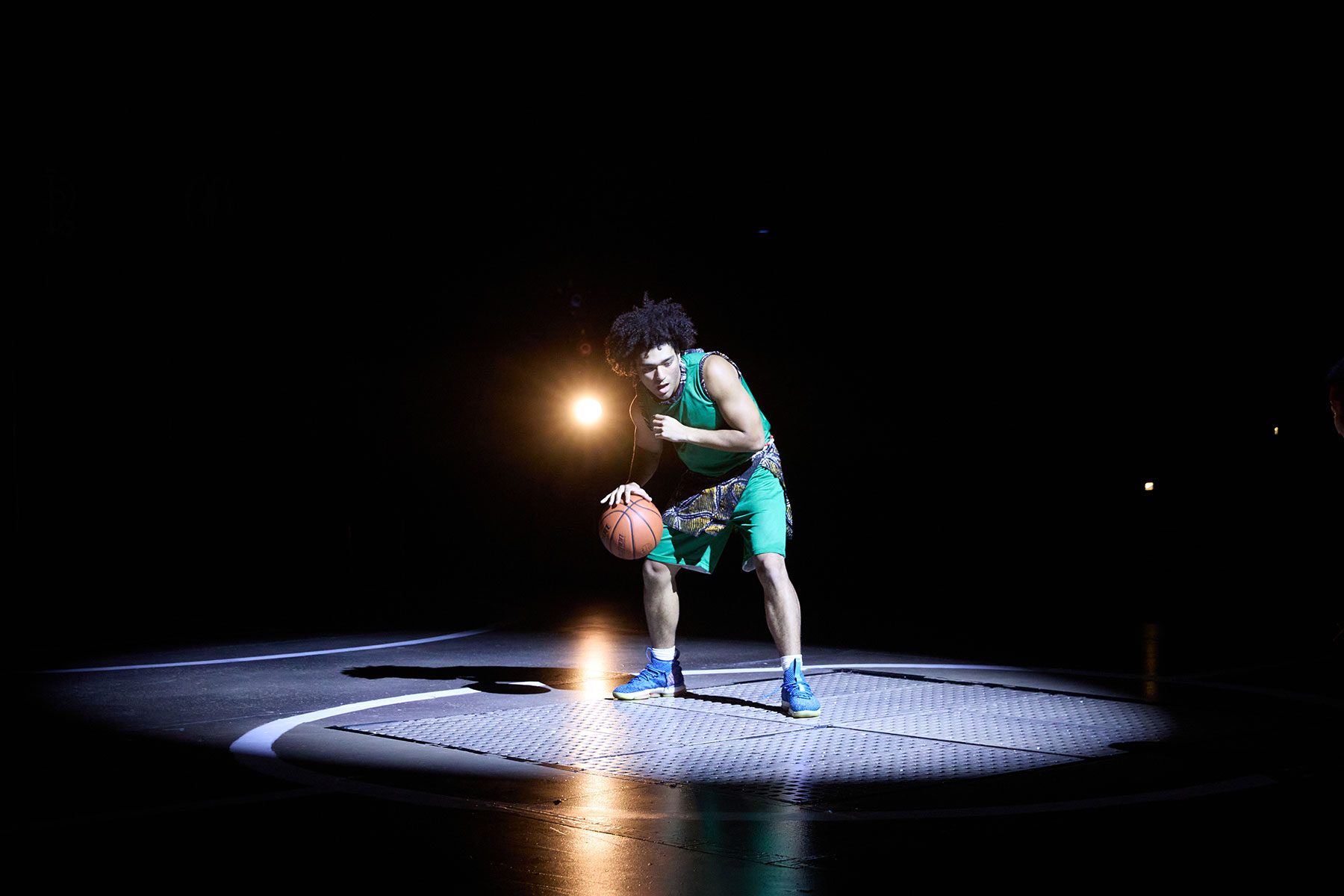 Basketball player in green outfit dribbles under spotlight on a dark court.