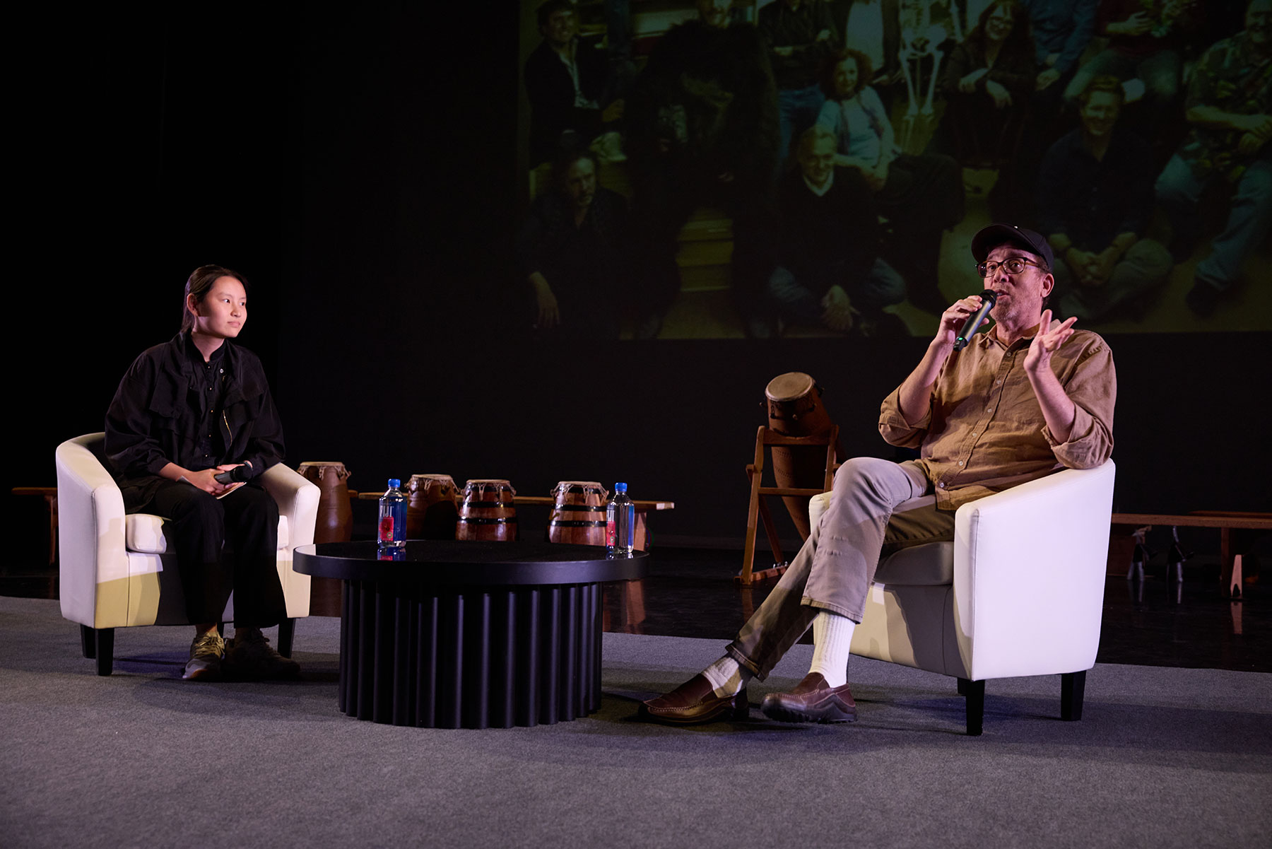 Two people seated on stage in white armchairs with microphones; drums and water bottles on a black table between them.