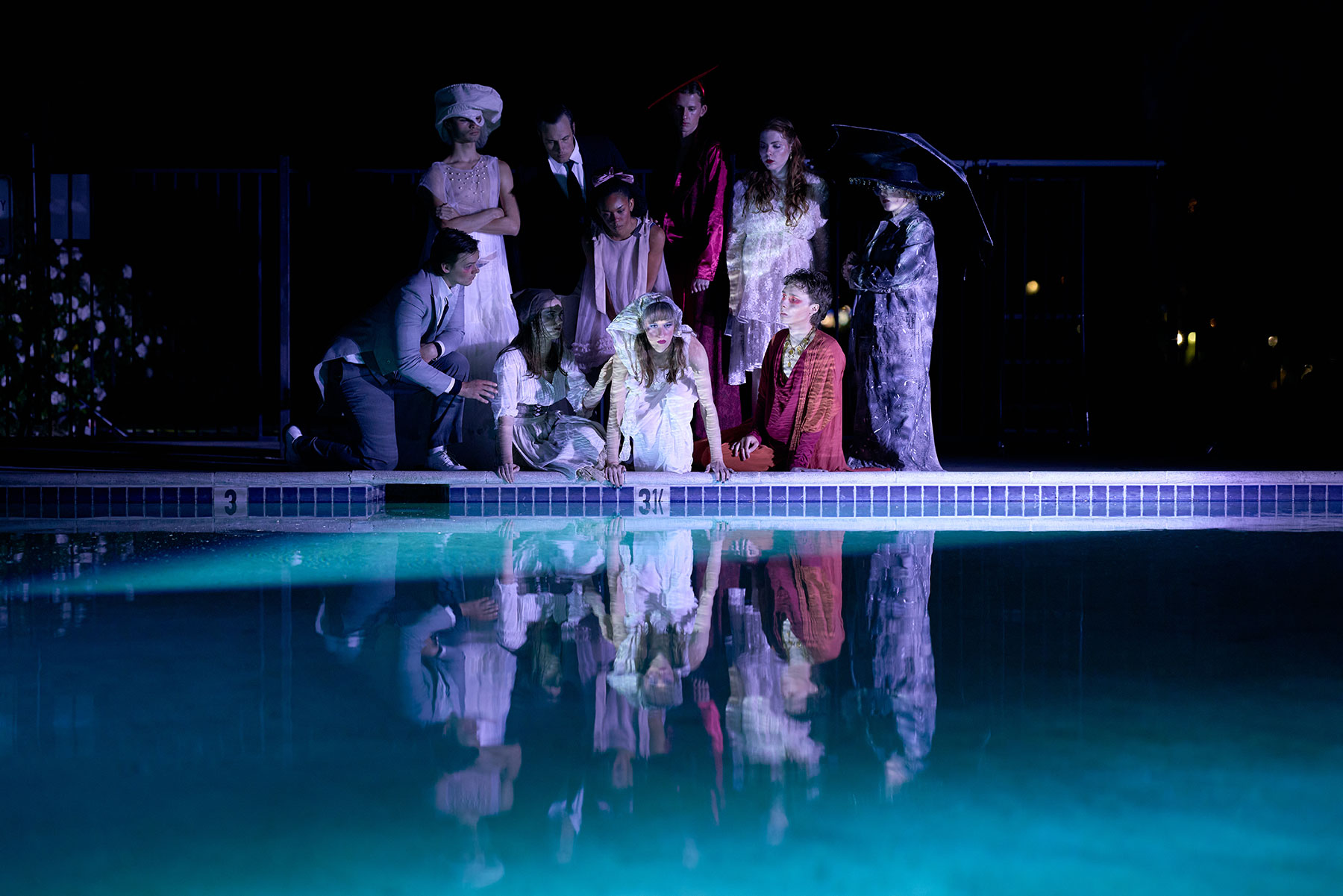 A group dressed in theatrical costumes stands by a dimly lit pool at night, with reflections in the water.
