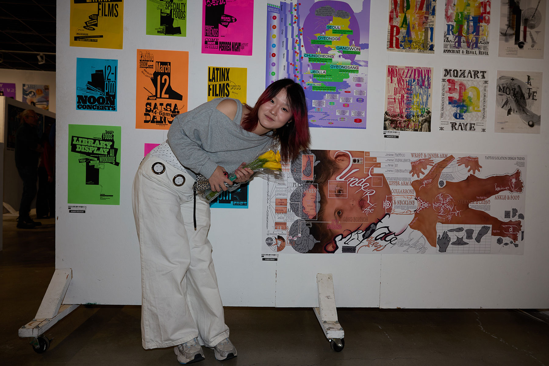 A person with red-tinted hair standing in front of a wall covered with colorful posters while holding yellow tulips.