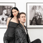 Two people, dressed in black, pose in front of erotic photos. She is standing while he sits.