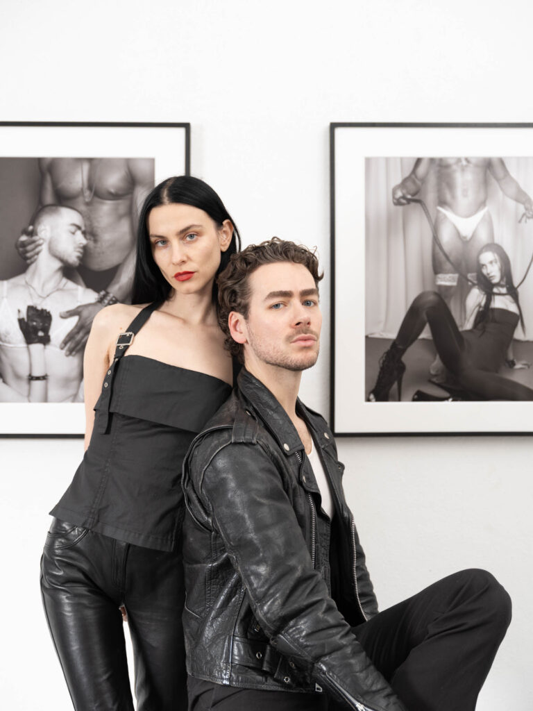 Two people, dressed in black, pose in front of erotic photos. She is standing while he sits.