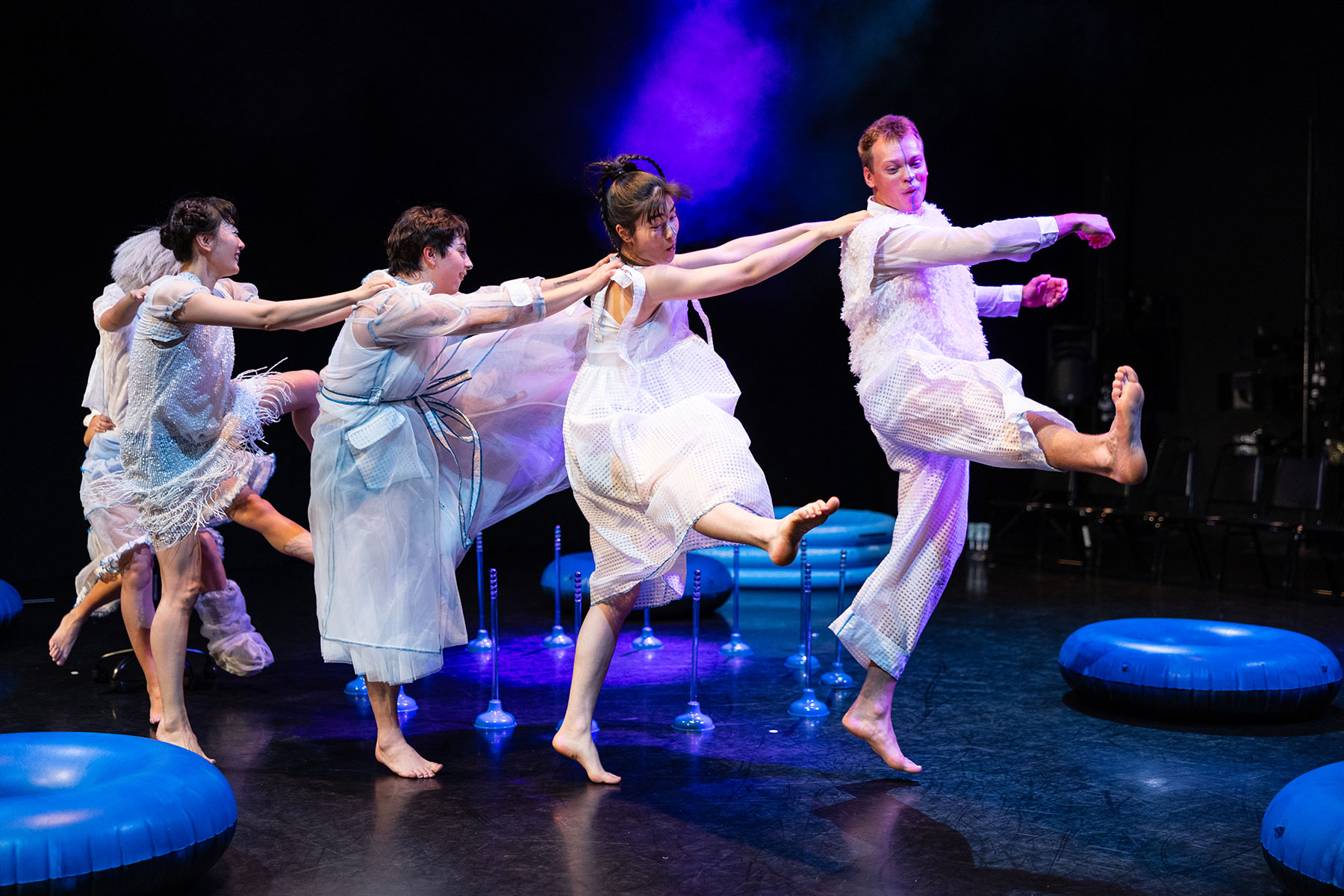 Five performers in white and blue costumes dancing energetically on a stage with blue inflatable props.