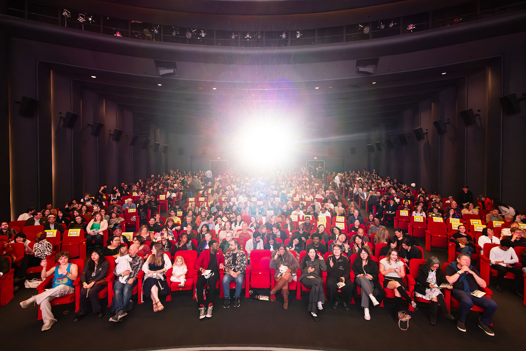 A large, crowded theater with people sitting in red seats, lit by a bright flash.