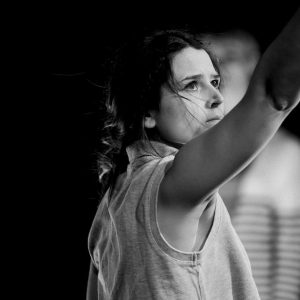 A black-and-white photo of a performer raising one arm while looking upward during a stage performance. Another figure appears out of focus in the background.