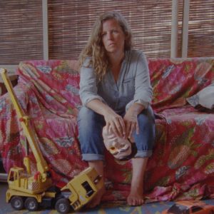 Image of a woman in denim sitting on a couch covered with a red-patterned cover. A small bulldozer lays at her feet.
