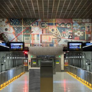 A view down the platform of a Los Angeles Metro D Line station featuring a large colorful geometric mural spanning the far wall above the tracks, with digital train signs and symmetrical station architecture in the foreground.