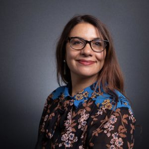 A woman with long, dark brown hair and glasses smiles against a dark gray background. She is wearing a brown floral blouse with a blue patterned collar and shoulder detail.