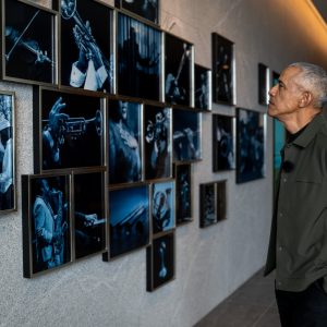 President Barack Obama stands in profile inside the Obama Presidential Center, viewing Carrie Mae Weems’ The Cool Blue Wind. The installation features a grid of blue-toned photographic images mounted along a wall, including scenes of jazz musicians and moments associated with Obama’s 2008 presidential campaign.