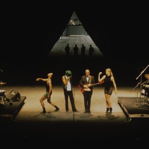 Four performers stand on stage, bookended by two drummers, with a triangle and their shadows at the back of the stage.