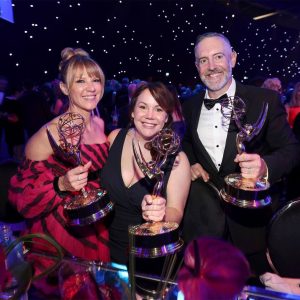 Three individuals in a theater smile at the camera, holding their Emmy Awards.