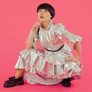 Atsuko Okatsuka crouching in a silver dress with hand on her chin against a pink backdrop.