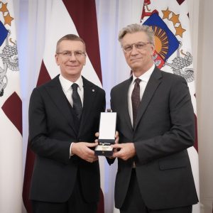 Two men in suits pose with a medal in front of official Latvian flags.