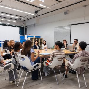 A group of people sitting around a table in a modern classroom, engaged in discussion.