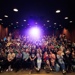 An at capacity crowd sits in a low-lit theater, backlit by the projector at the back of the room.