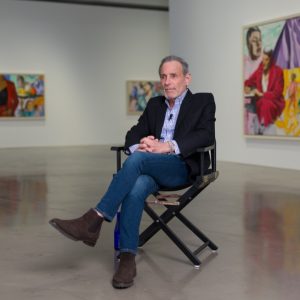 A man sits on a director's chair in the middle of a gallery.