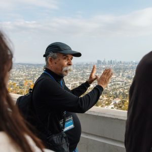 Harry Gamboa Jr. wearing a ball cap frames the skyline of LA with his hands while talking to a class of students.