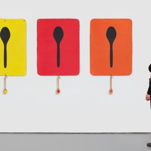 A woman in black looks at three artworks of spoons with spoons hanging from the bottom of the artworks.