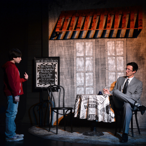 Man in a gray suit doing crossword puzzle outside a cafe looks at a young boy in a red hoodie.