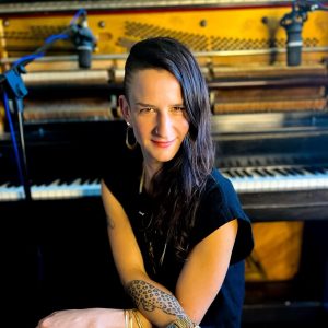 A woman in black sits at a piano, turned around and facing the camera