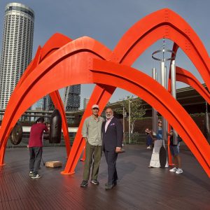 Two men posing under a massive, bright red arched metal sculpture, resembling a spider, situated on an outdoor wooden deck in front of a modern skyscraper and other city buildings. The sculpture has large gongs and wind chimes hanging from its frame.