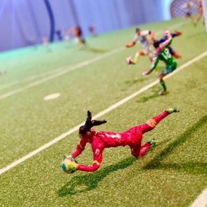 Miniature soccer scene from Fútbol Is Life featuring a goalkeeper in a red uniform diving to block a shot near the goal, with other players visible in the background on a detailed green pitch.