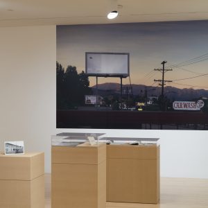 Gallery setting with a large photograph of a billboard and skyline at dusk, alongside two display pedestals. Transcribed Text: CAR WASH COIN OP Ralphs