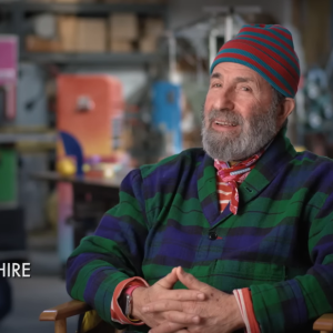 Peter Shire, smiling, sits in colorful studio with hands clasped