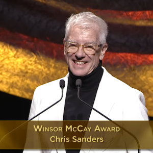 A screenshot of a smiling man at a podium. He is wearing a black turtleneck, a cream blazer and glasses. The overlay reads, "Winsor McCay Award / Chris Sanders."