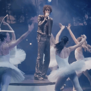 A singer in a studded leather outfit performs on stage surrounded by ballet dancers.