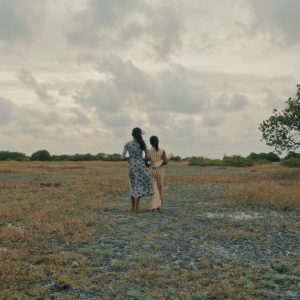 Two women in dresses walk away from the camera across a dry, grassy field toward a large green tree on the right under a cloudy, overcast sky.